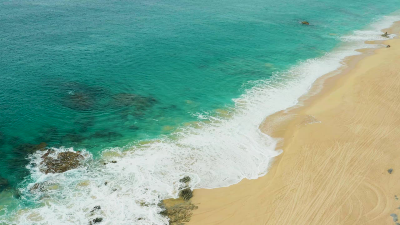 Aerial frame of Laguna Beach Los Cabos showing turquoise water, white foam, dark rocks, pale sand, beach tracks, and no visible people at the start of the clip.