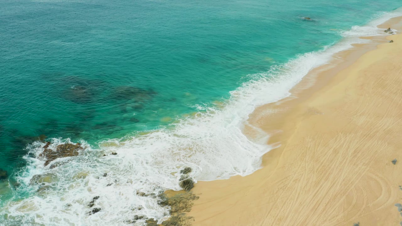 Aerial frame of Laguna Beach Los Cabos where the foam line pushes farther across the sand, with rocks at the lower left and no visible people.