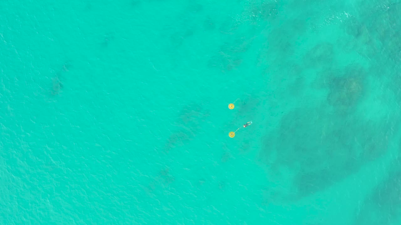 Top-down view of clear turquoise water at Laguna Beach Los Cabos with two yellow buoys near a distant swimmer and faint darker seabed patches below.