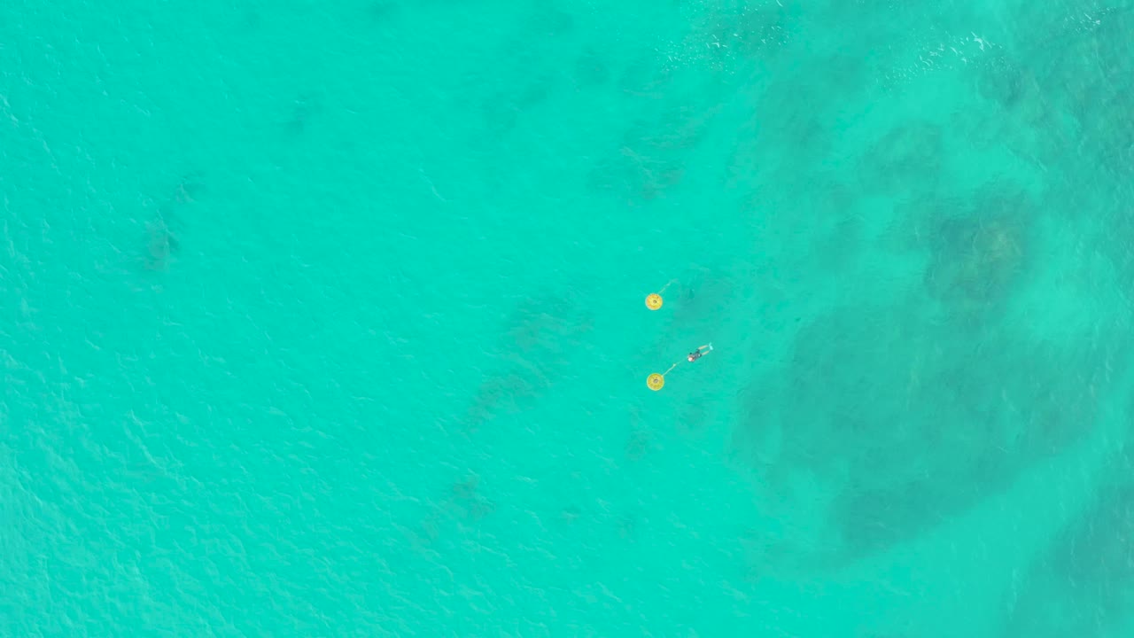Aerial turquoise ocean with yellow buoys and a small swimmer near center