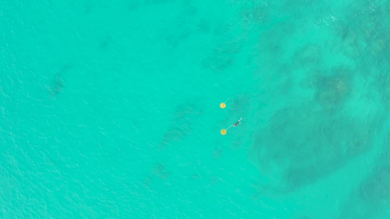 Clear blue-green water at Laguna Beach Los Cabos fills the frame as two yellow buoys sit to the left of a distant swimmer above mottled seabed tones.
