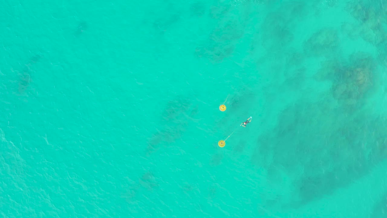 Overhead view in Laguna Beach Los Cabos, Mexico, showing a small snorkeler just right of two yellow buoys over clear turquoise water with darker submerged formations.