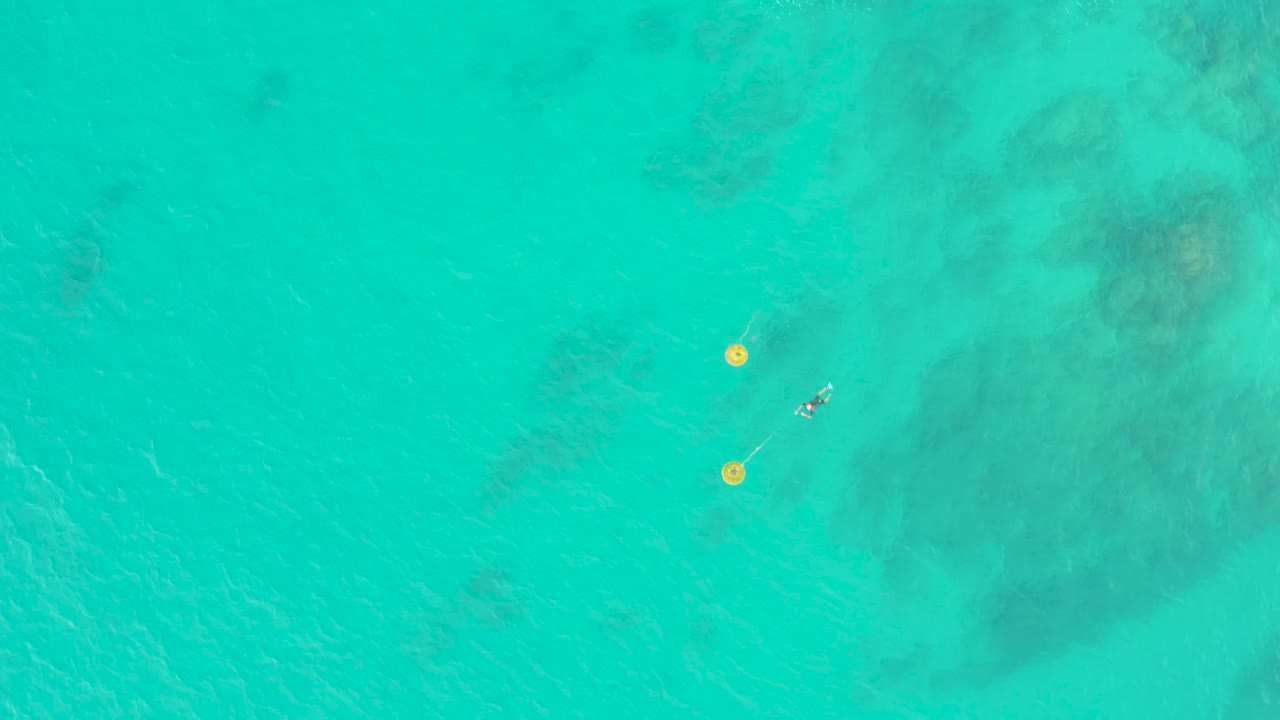 Aerial frame from Laguna Beach Los Cabos, Mexico, with the snorkeler slightly separated from the two yellow buoys and faint coral or rock shapes visible below the water.