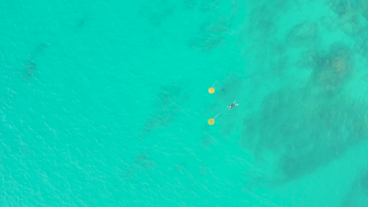 Laguna Beach Los Cabos aerial frame with two yellow buoys low in the composition and a small snorkeler nearby over transparent blue-green water.