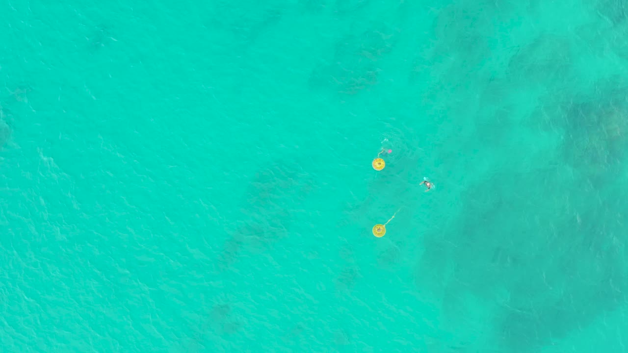 Top-down view of clear turquoise water at Laguna Beach Los Cabos with two yellow buoys and a small swimmer near darker underwater patches.
