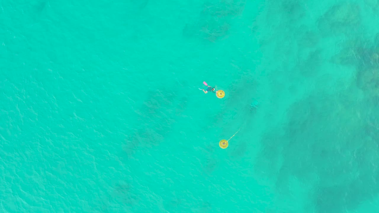 Overhead view of clear turquoise water at Laguna Beach Los Cabos, with two yellow floats and small swimmers near the upper part of the frame.