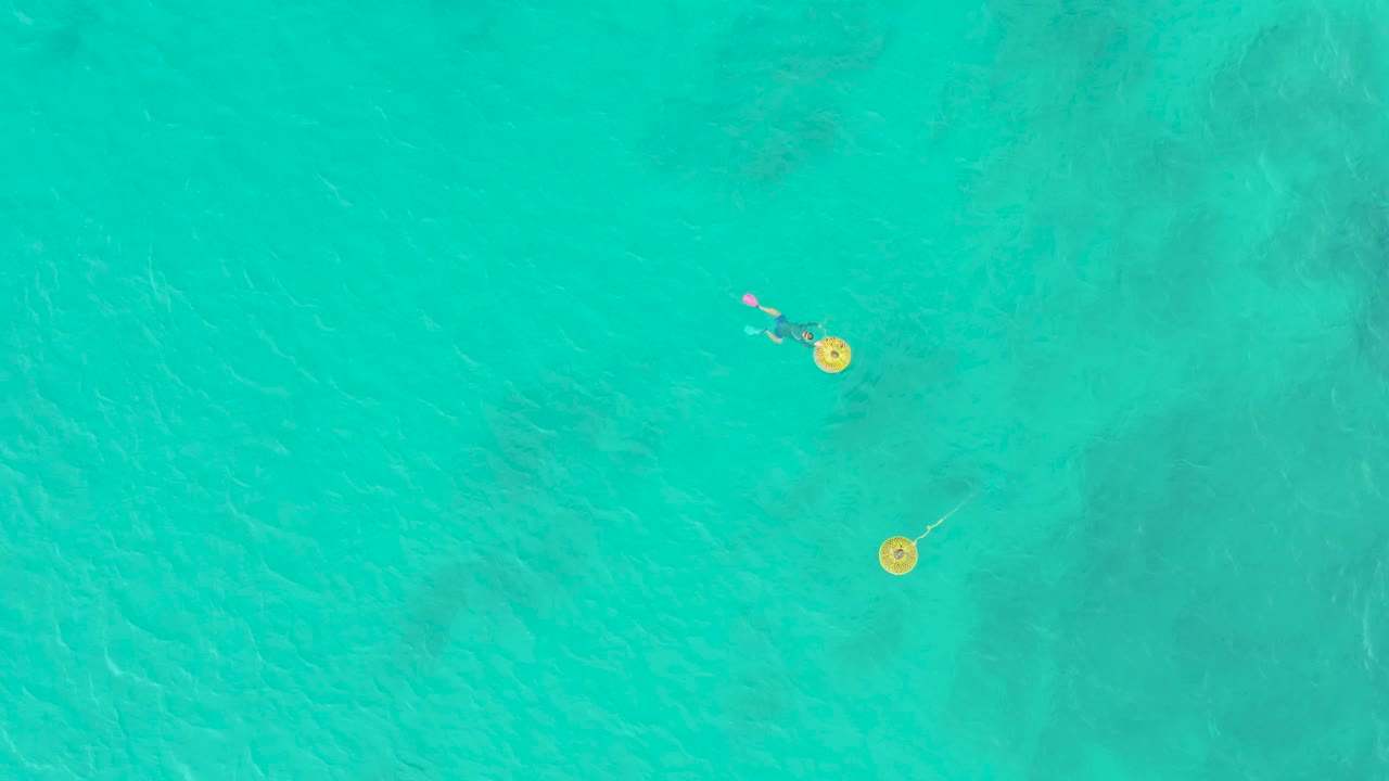 Overhead view at Laguna Beach Los Cabos showing a small swimmer with a pink floatie near two yellow buoys in clear turquoise water.
