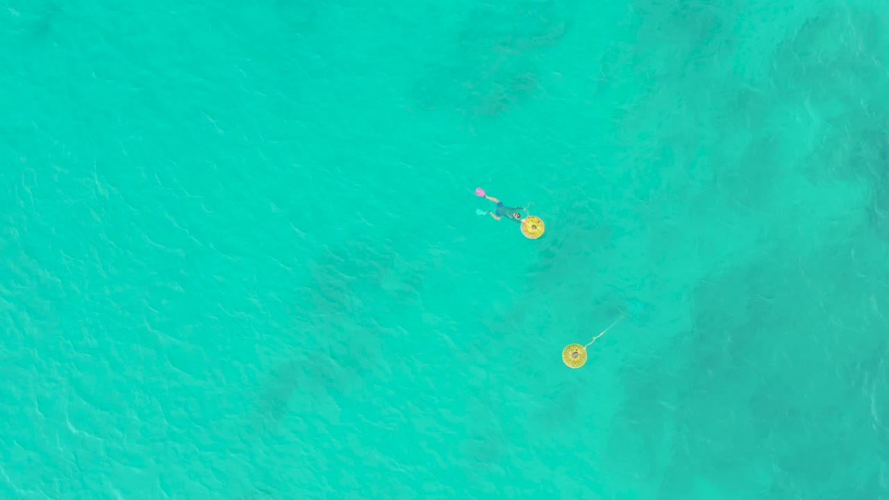 Swimmer and yellow buoys seen from directly above turquoise water.