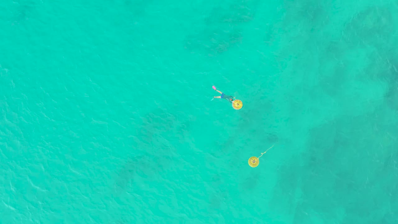 Top-down Laguna Beach Los Cabos frame showing the swimmer near the top of the composition, with two yellow buoys spaced diagonally below.