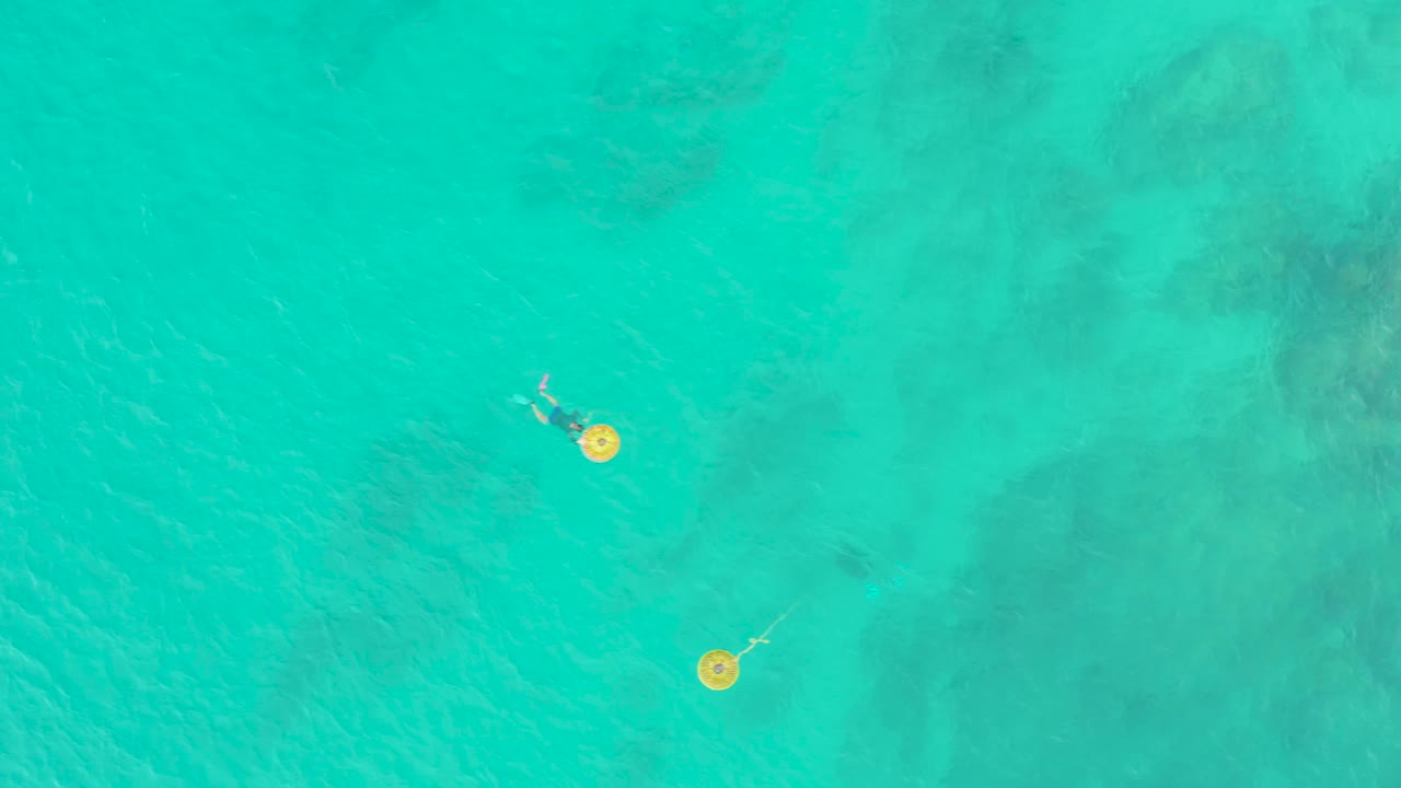Overhead frame at Laguna Beach Los Cabos shows a person near a yellow circular float in clear turquoise water with faint seabed patterns.