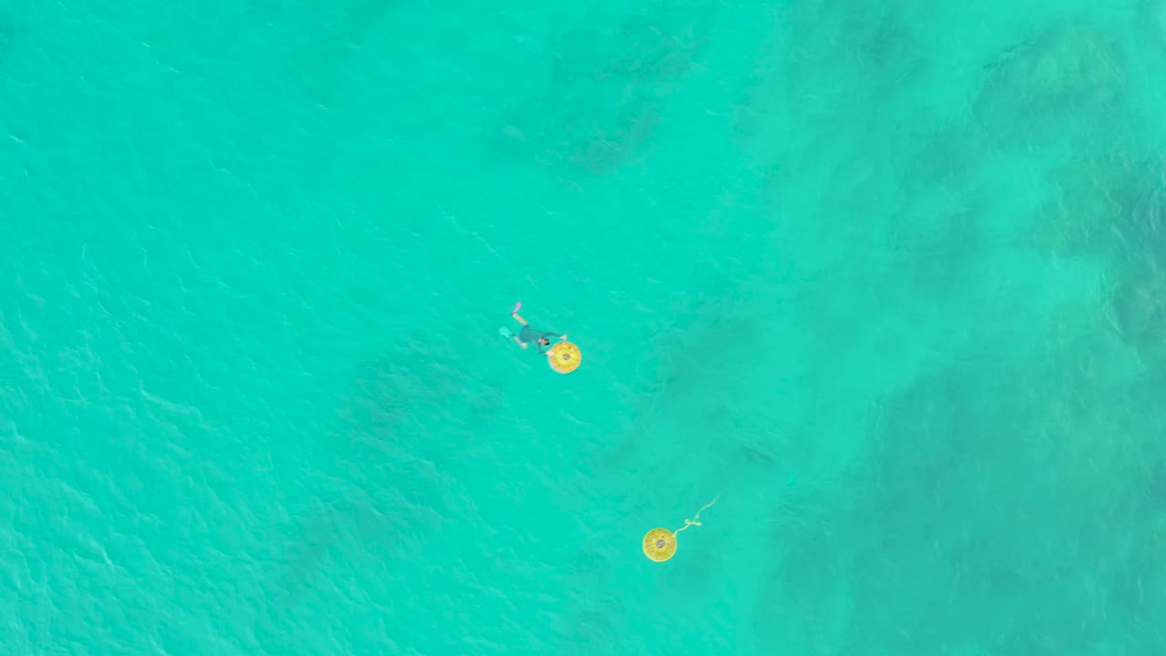 At 0:02, the yellow float and swimmer remain near the upper portion of an overhead turquoise ocean frame with soft darker seabed areas below.