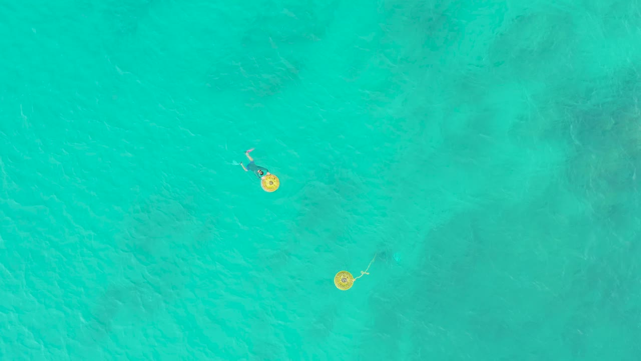 Overhead view at Laguna Beach Los Cabos of two people on yellow float rings separated across clear turquoise water with faint seabed patches.