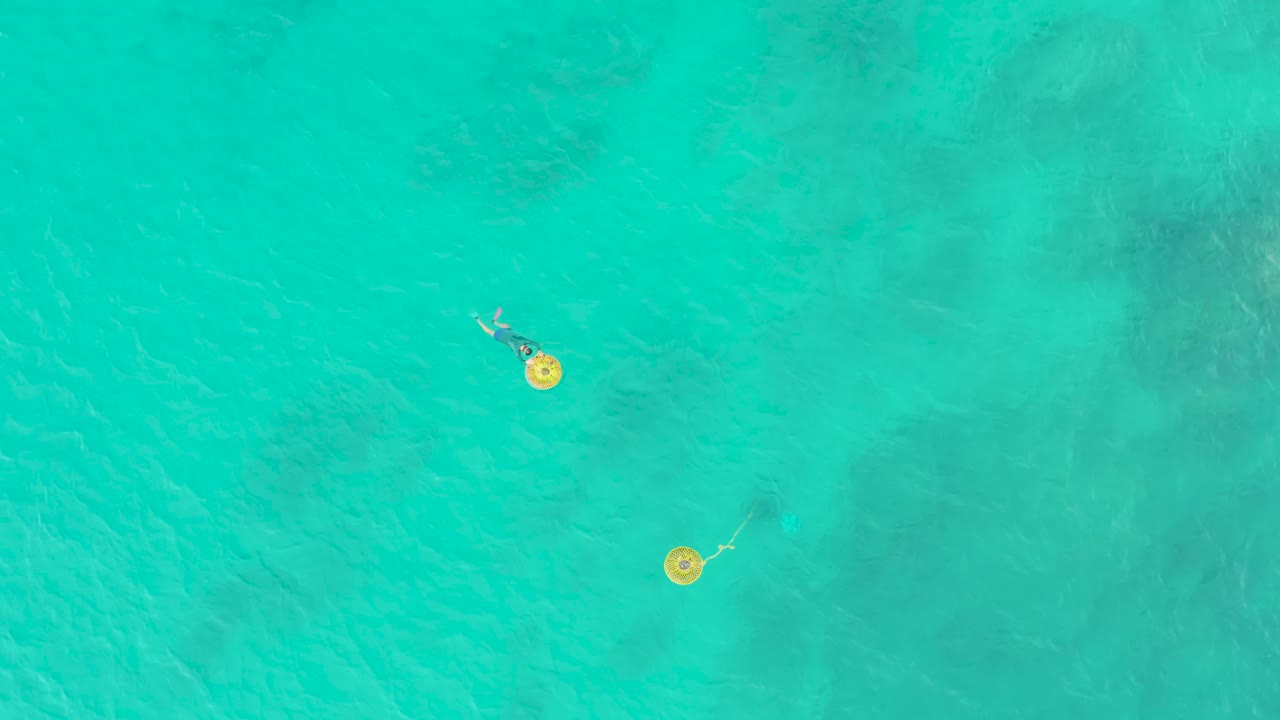 Direct overhead view in Laguna Beach Los Cabos showing two people floating on yellow rings over rippled turquoise water with muted seabed areas.