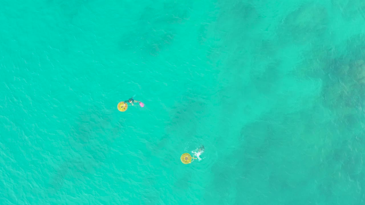 At 0:00 in Laguna Beach Los Cabos, two people float far apart on yellow rings over clear turquoise ocean with darker seabed patches beneath.