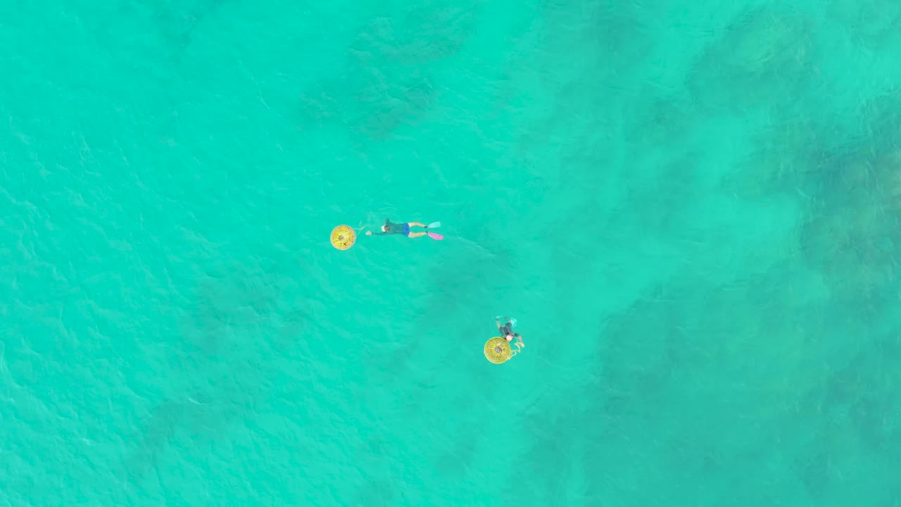 Aerial frame of two people in bright turquoise sea at Laguna Beach Los Cabos, with yellow floats standing out against darker underwater patches.