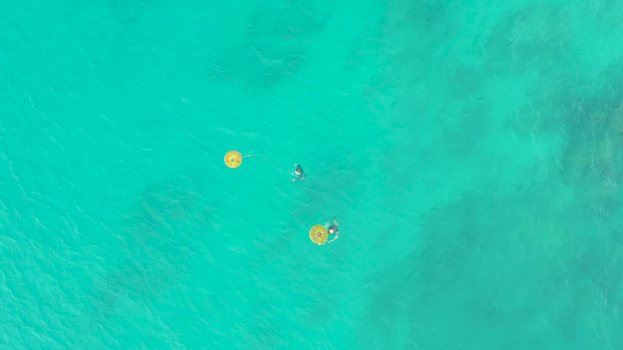 Overhead view at Laguna Beach Los Cabos of two visible swimmers spaced apart near yellow float rings in clear turquoise ocean water.