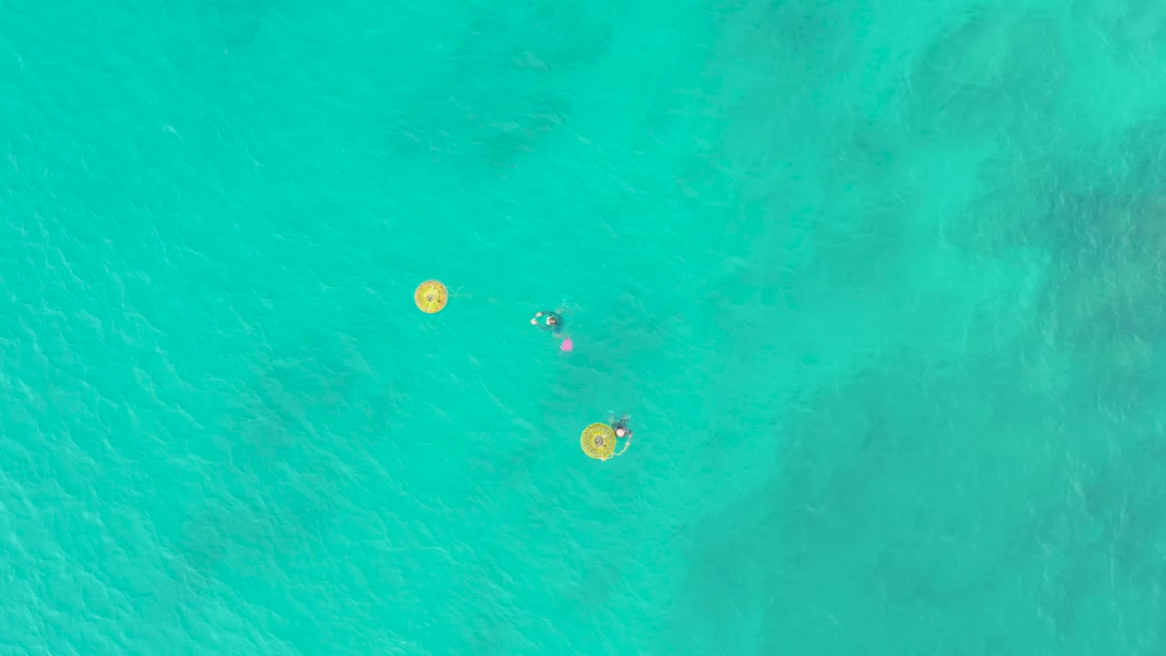 Overhead view of two visible swimmers in Laguna Beach Los Cabos water, with one swimmer closer to a yellow ring and another farther across the turquoise surface.