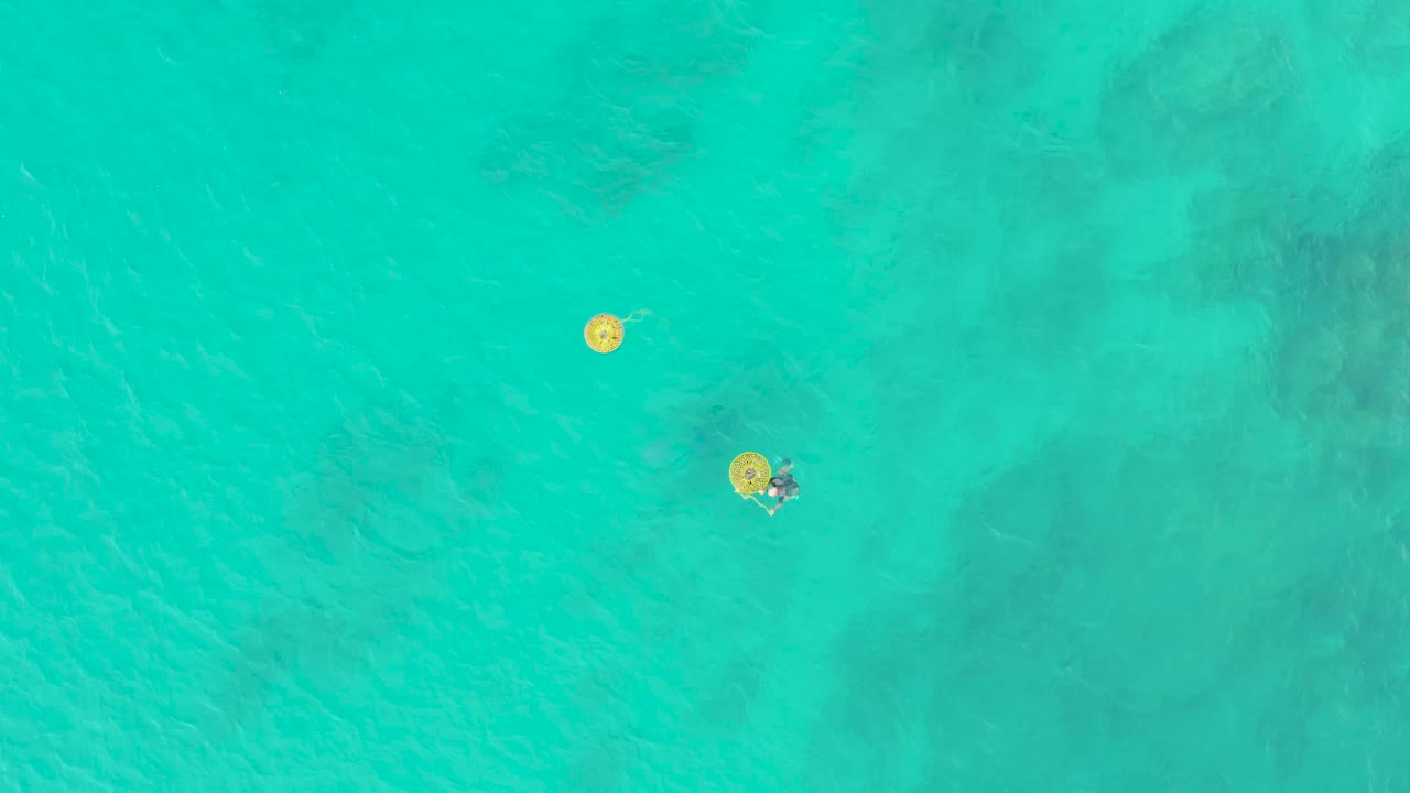 At 0:02, the top-down view shows the upper yellow float and a swimmer near the lower yellow float against clear turquoise water and faint seabed shading.