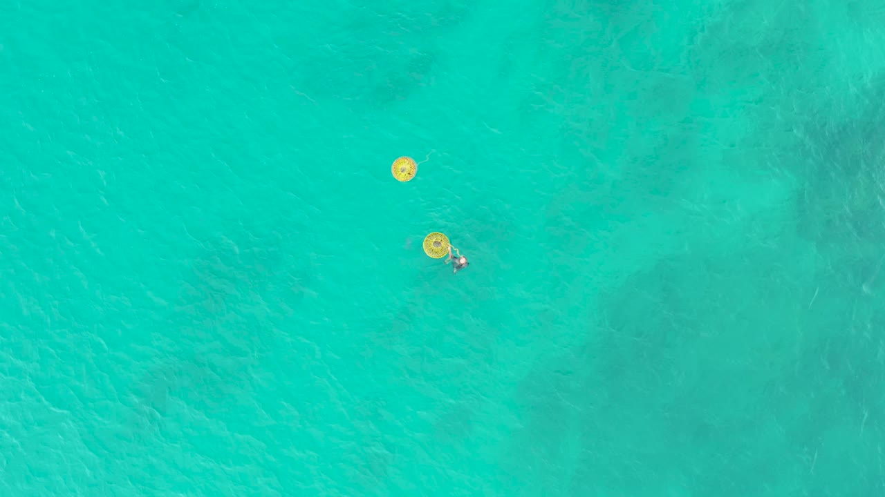 Overhead view at Laguna Beach Los Cabos shows clear turquoise water, two yellow buoys, and a person floating beside the lower buoy near the start of the clip.