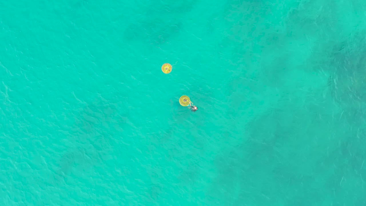 At 0:02, the overhead frame shows two yellow floats in clear water, with the person still visible beside the lower float and faint ripples crossing the surface.