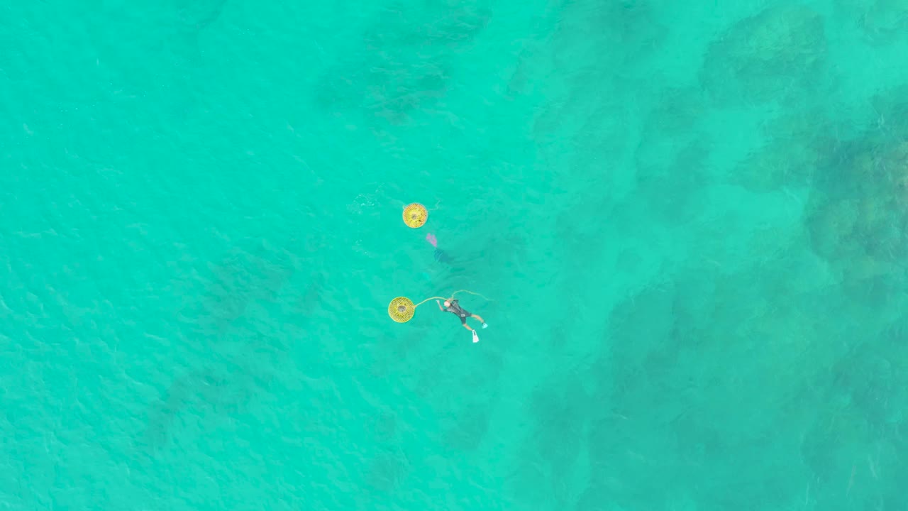 At 0:00, an overhead view at Laguna Beach Los Cabos shows two swimmers beside separate yellow buoys in clear turquoise water with darker seabed patches below.