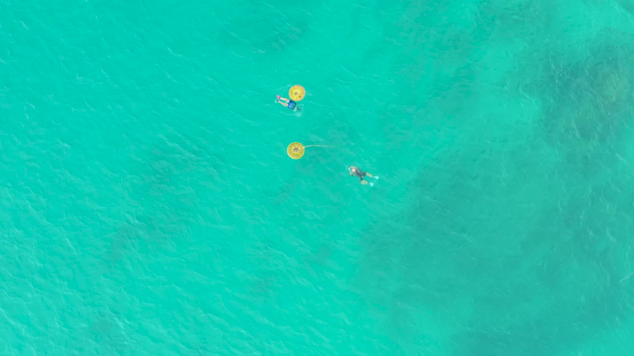 High overhead view at Laguna Beach Los Cabos showing two swimmers separated in clear turquoise water, with two yellow buoys near the upper left area of the frame.