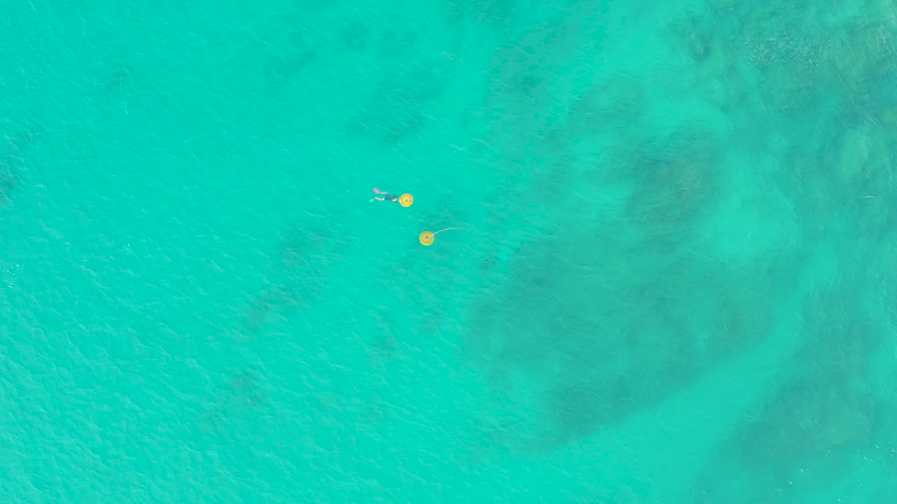 Overhead turquoise water with a swimmer near two yellow buoys.