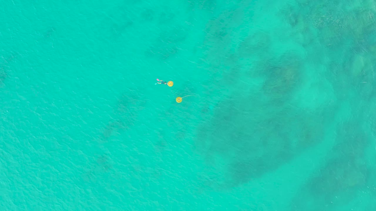 At 0:01, the Laguna Beach Los Cabos aerial frame keeps the swimmer close to the two yellow buoys over pale turquoise water and darker seabed patches.