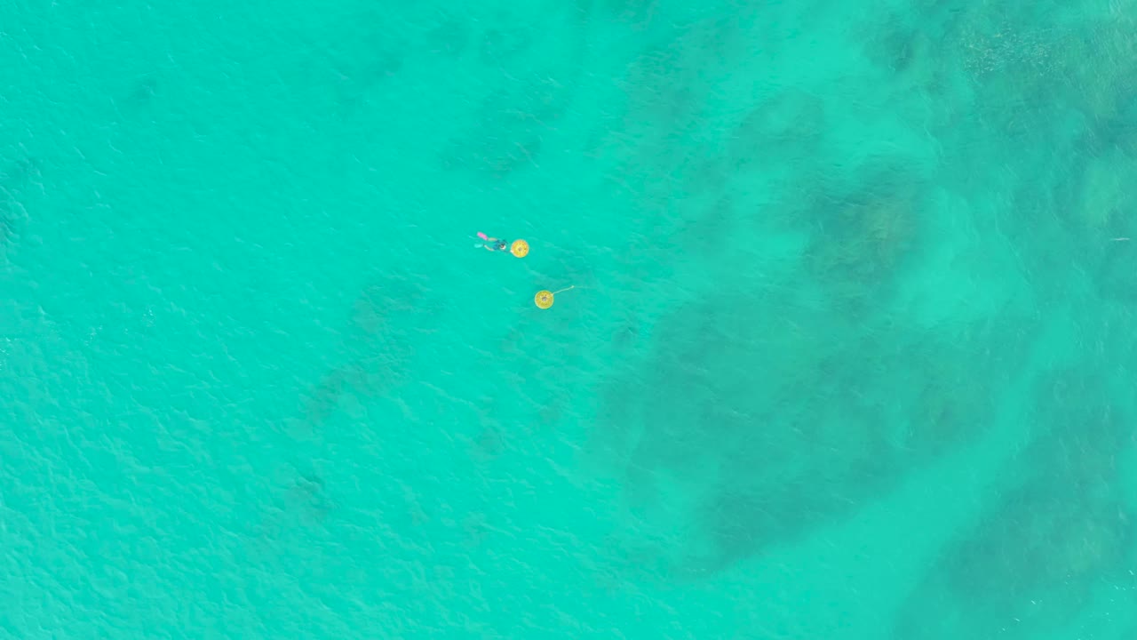 At 0:02, the overhead view shows the swimmer slightly offset from the yellow buoys, surrounded by clear turquoise water with muted seabed color below.