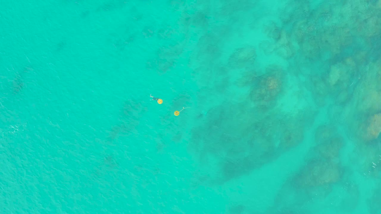 Top-down view of clear turquoise water at Laguna Beach Los Cabos with two yellow floats and tiny swimmers near darker underwater rock patches.