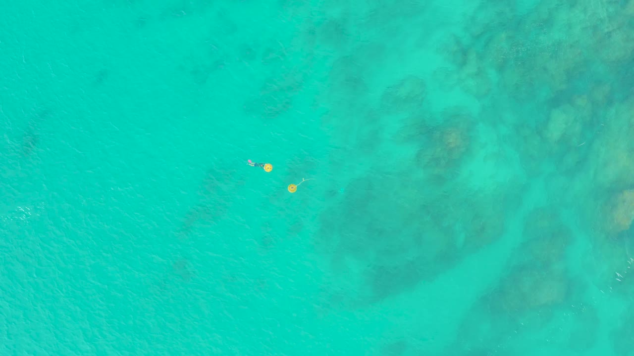 Two yellow floats and tiny swimmers appear near the upper-left area of a broad turquoise frame, with darker underwater rocks spreading across the right side.