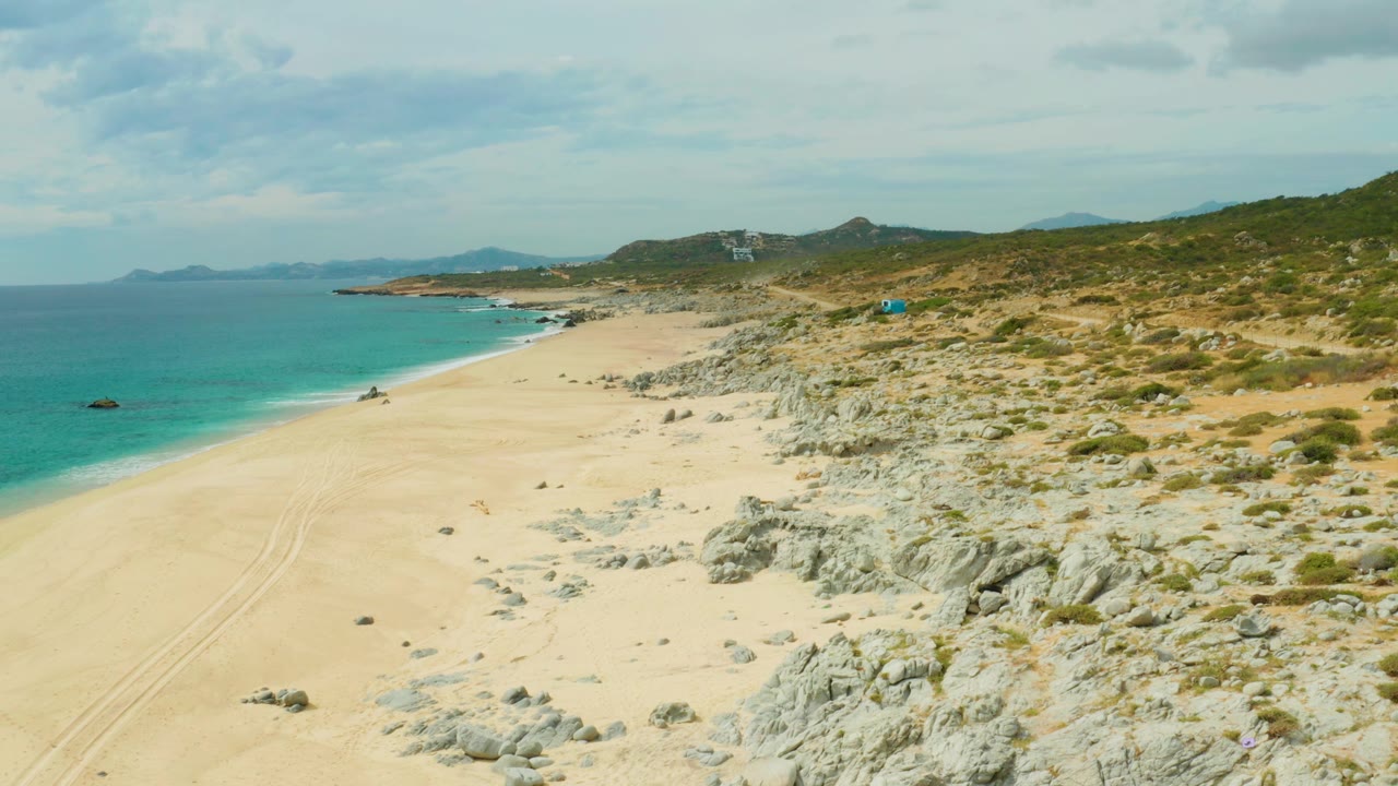 Laguna Beach Los Cabos coastline seen from above, with the sandy beach stretching toward distant hills, turquoise water along the left edge, rocks and scrub on the right, a small boat near shore, and no visible people.