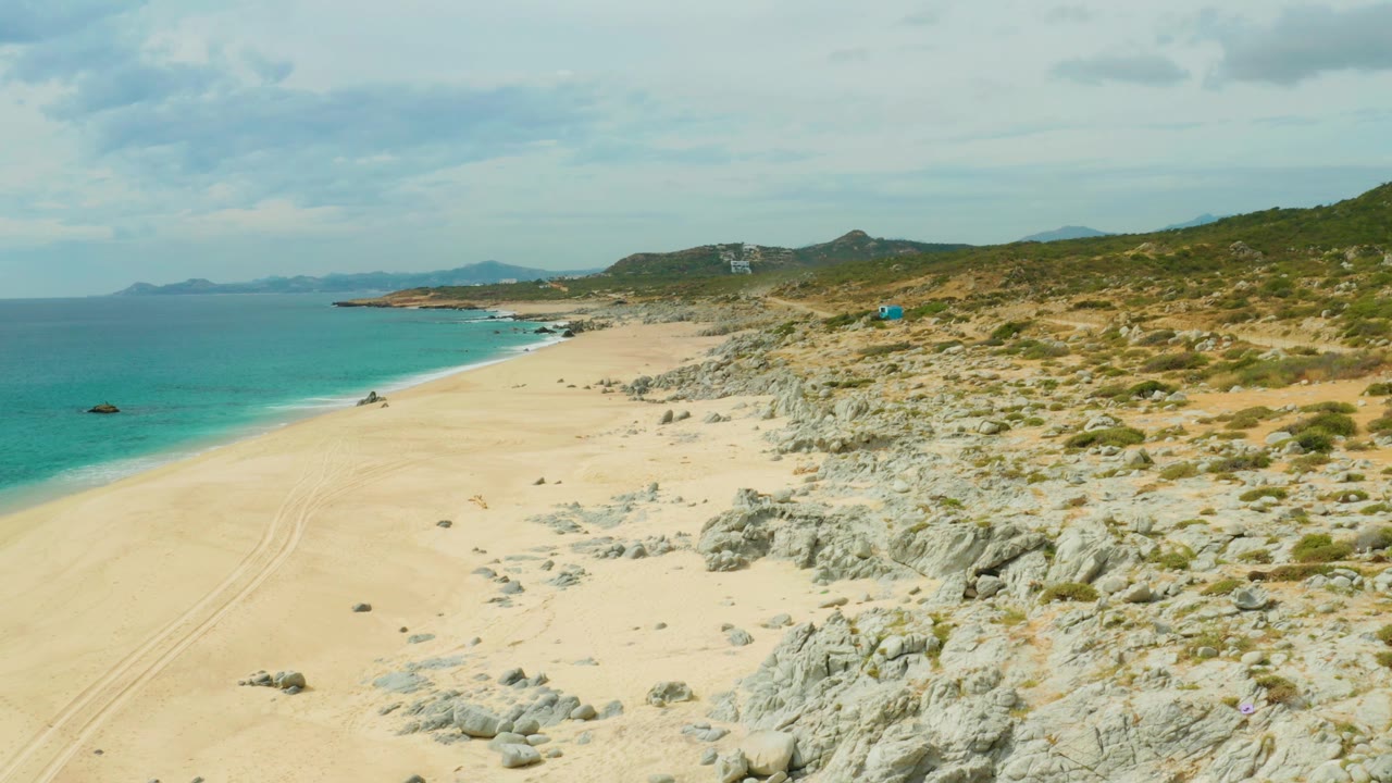 A high wide view over Laguna Beach Los Cabos shows empty sand, turquoise ocean, rocky dune slope, distant headlands on the horizon, scattered clouds, and no visible people.