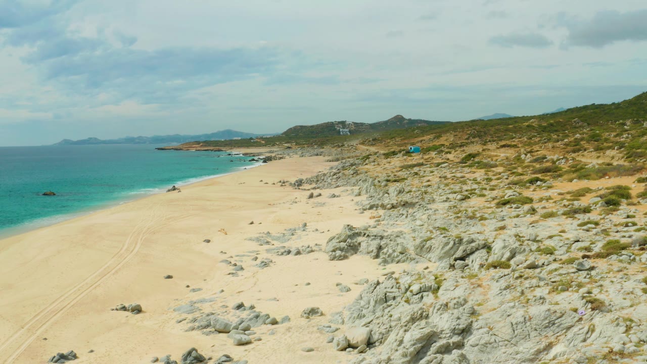 Beach and rocky dune coastline with blue-green water and clouded sky.