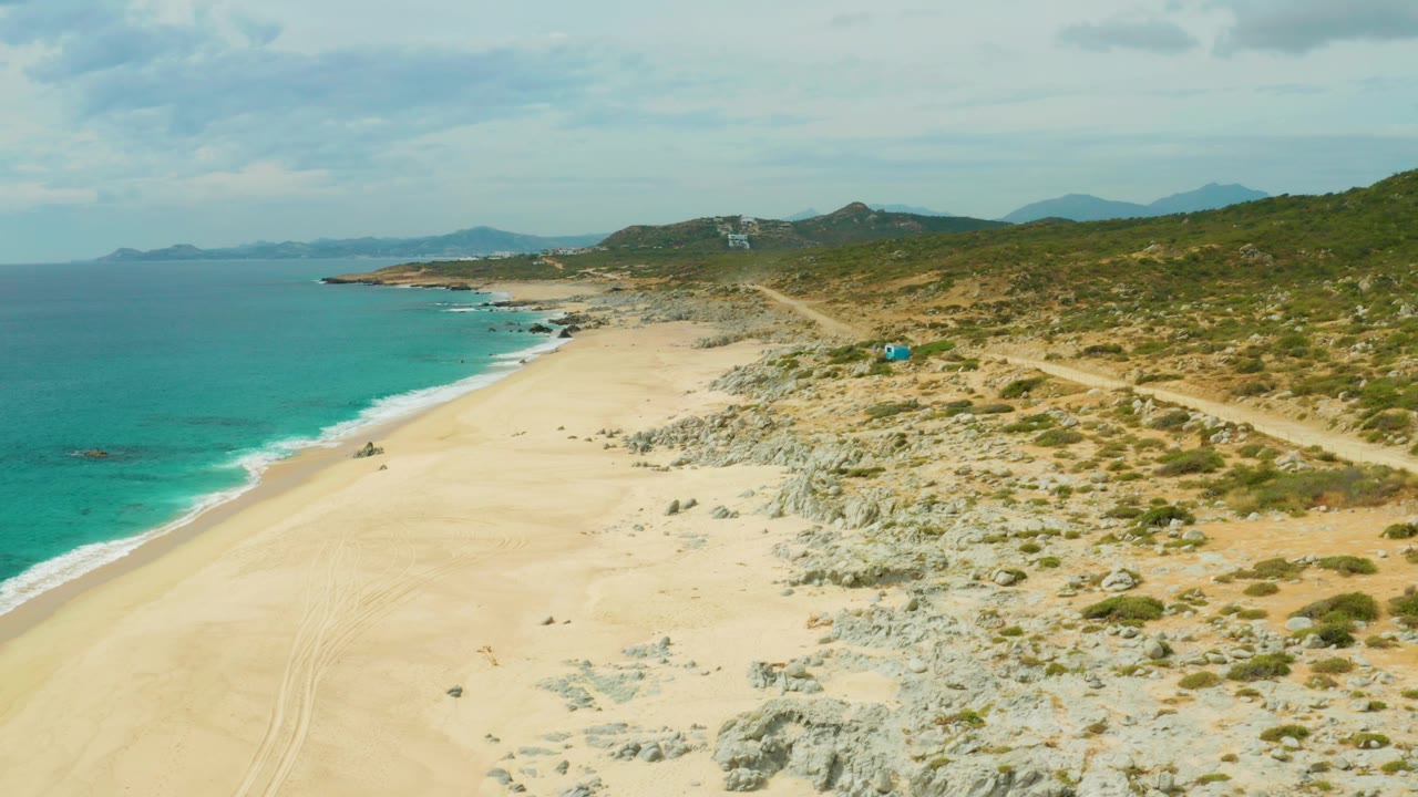 At 0:00, an aerial view over Laguna Beach Los Cabos shows pale sand, turquoise surf, rocky dune terrain, a small blue vehicle on the right-side dirt path, distant hills, and no visible people.