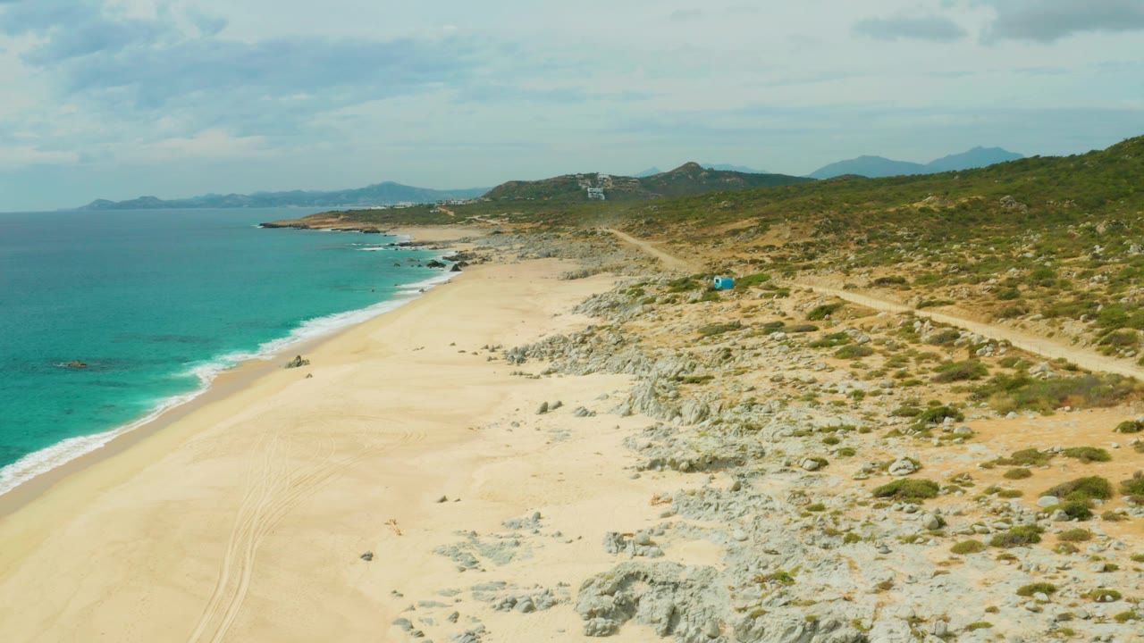 At 0:01, the drone view remains high over Laguna Beach Los Cabos, with the shoreline angled along the left, the blue vehicle slightly farther along the sandy path, rocky scrub on the right, and no visible people.