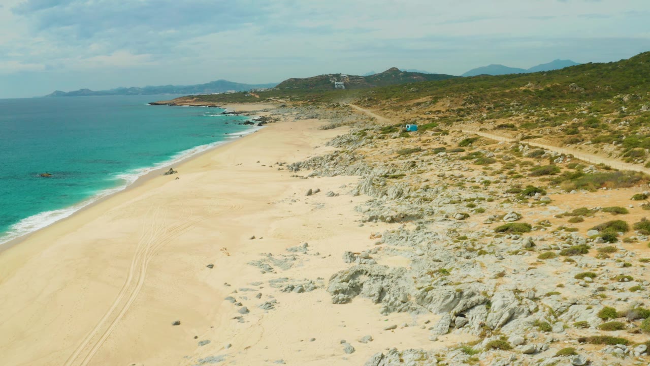 Aerial beach frame with tire tracks in sand, turquoise ocean, and a blue vehicle near scrubby rocks.