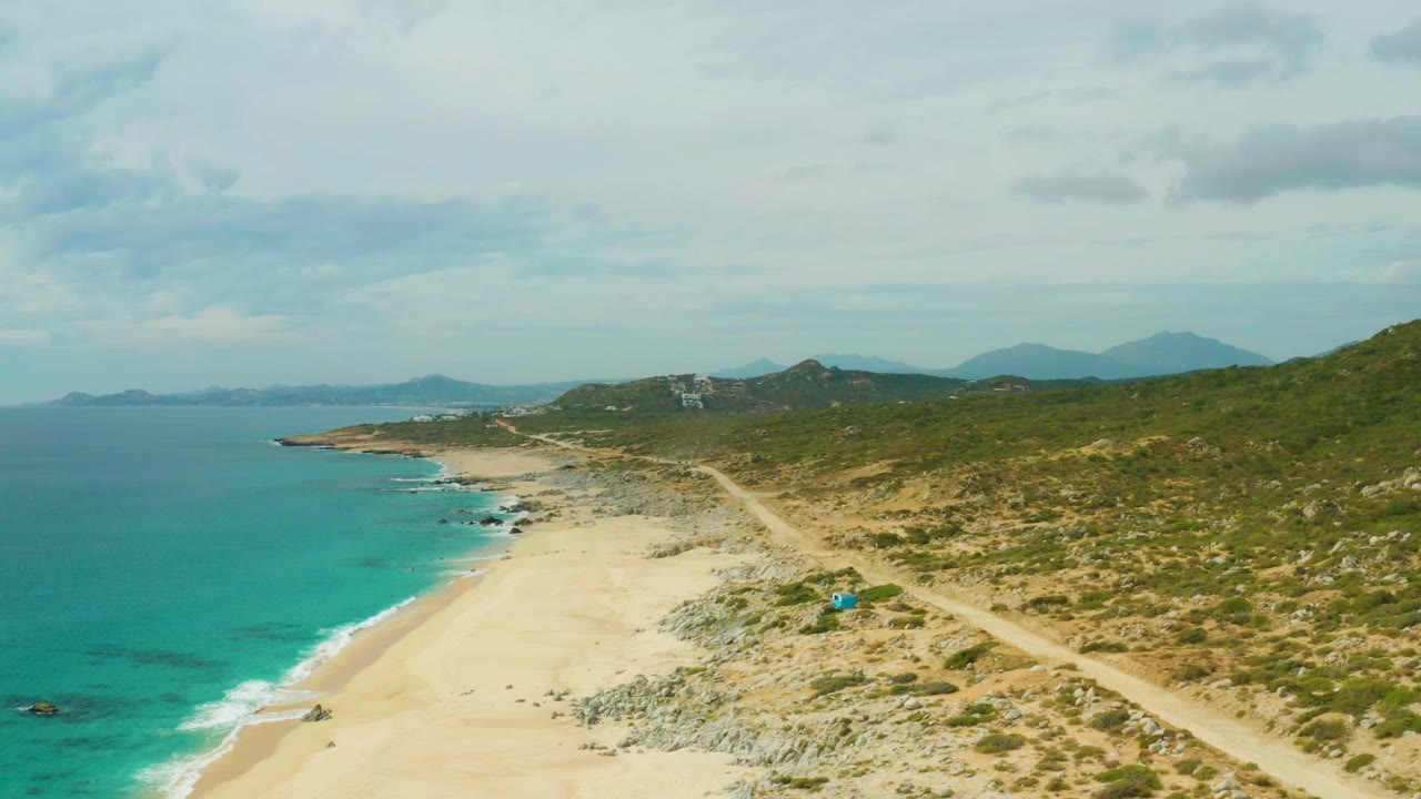 At 0:00, an elevated view over Laguna Beach Los Cabos shows turquoise water, pale sand, a rocky beach edge, green hills, a dirt road, distant mountains, and no visible people.