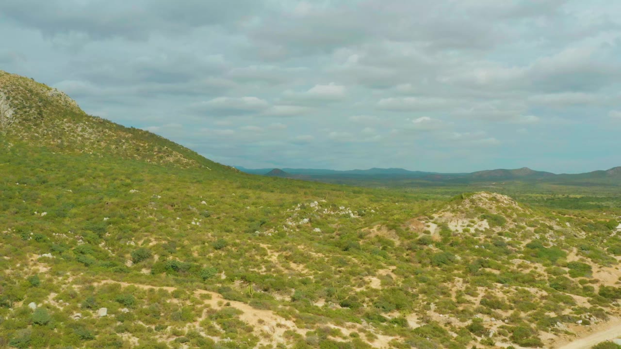 A green rocky hillside fills the left side of the Laguna Beach Los Cabos landscape, with open scrubland, far hills, cloudy sky, and no visible people.