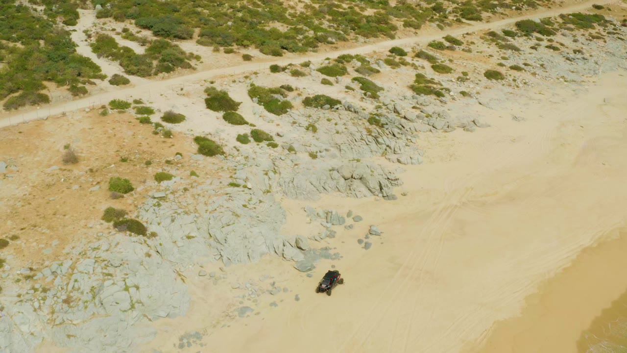 Drone view over Laguna Beach Los Cabos with the dark vehicle lower in frame, pale sand through the center, gray rocks above it, scattered shrubs, and no visible people.