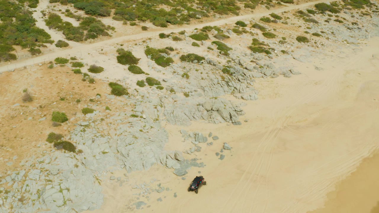 High aerial frame in Laguna Beach Los Cabos showing a dark vehicle near the lower right, broad pale sand to the side, gray rock formations, sparse shrubs, and no visible people.