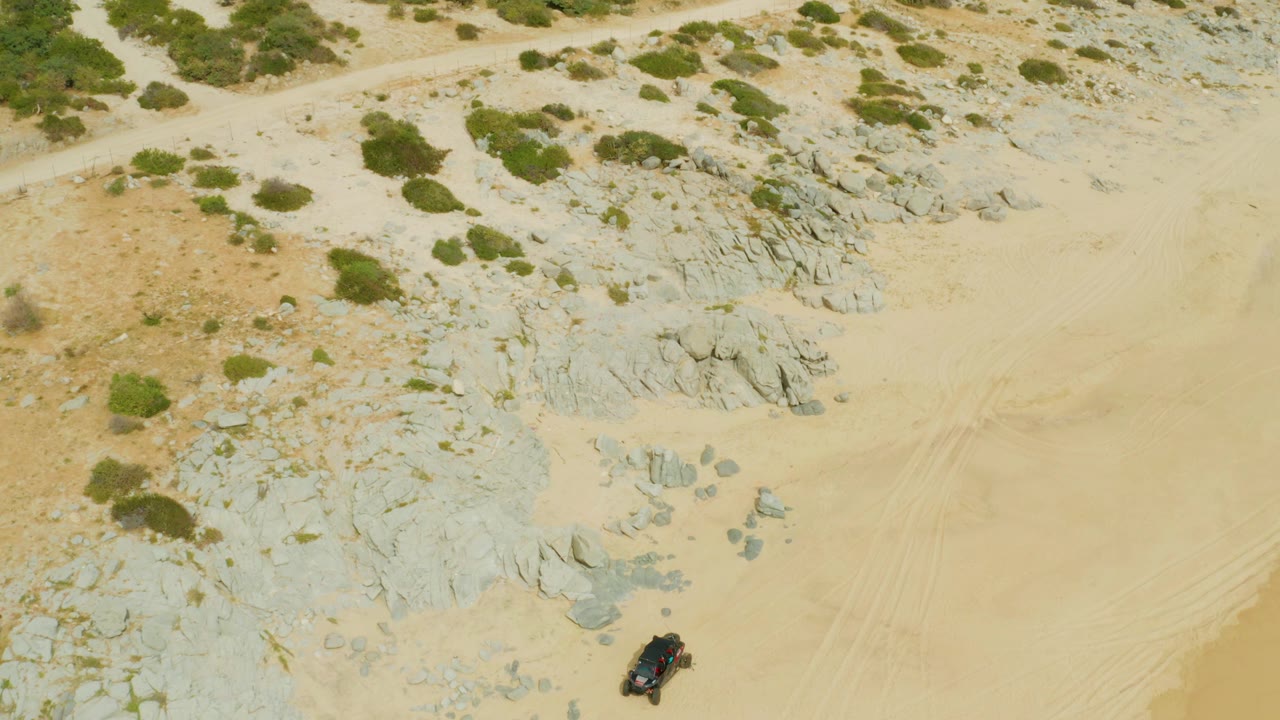 Vehicle at the lower edge of a sandy desert frame with shrubs and rocks above