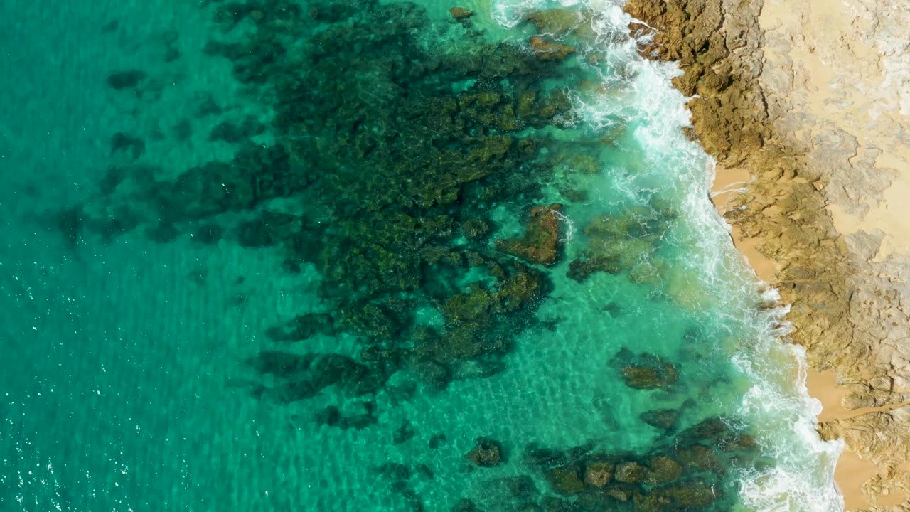 Clear turquoise water shows broad dark rock patches beneath the surface as foam traces the tan coast at Laguna Beach Los Cabos, with no visible people.