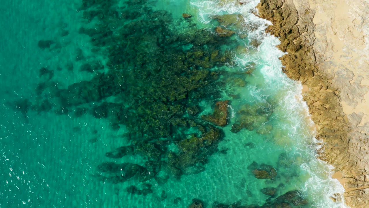 White foam breaks across the upper-right rocky shore while shallow turquoise water and dark reef shapes fill the left side at Laguna Beach Los Cabos, with no visible people.