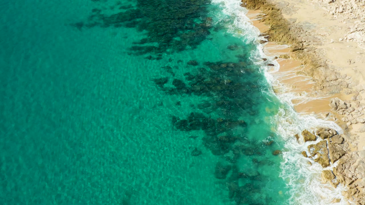 Aerial frame from Laguna Beach Los Cabos showing turquoise water, dark underwater rocks, white surf, pale sand, and no visible people at the start of the clip.