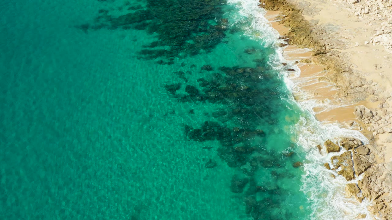 Clear turquoise ocean with dark rocks under the surface beside a sandy rock coast.