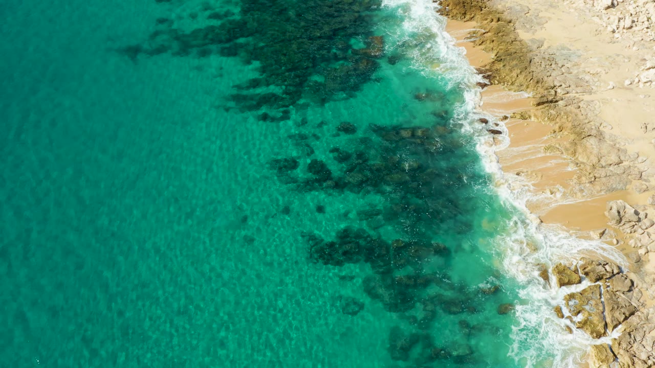 Aerial view over Laguna Beach Los Cabos where white foam pushes into the rocky right edge while turquoise water covers dark underwater formations, with no visible people.