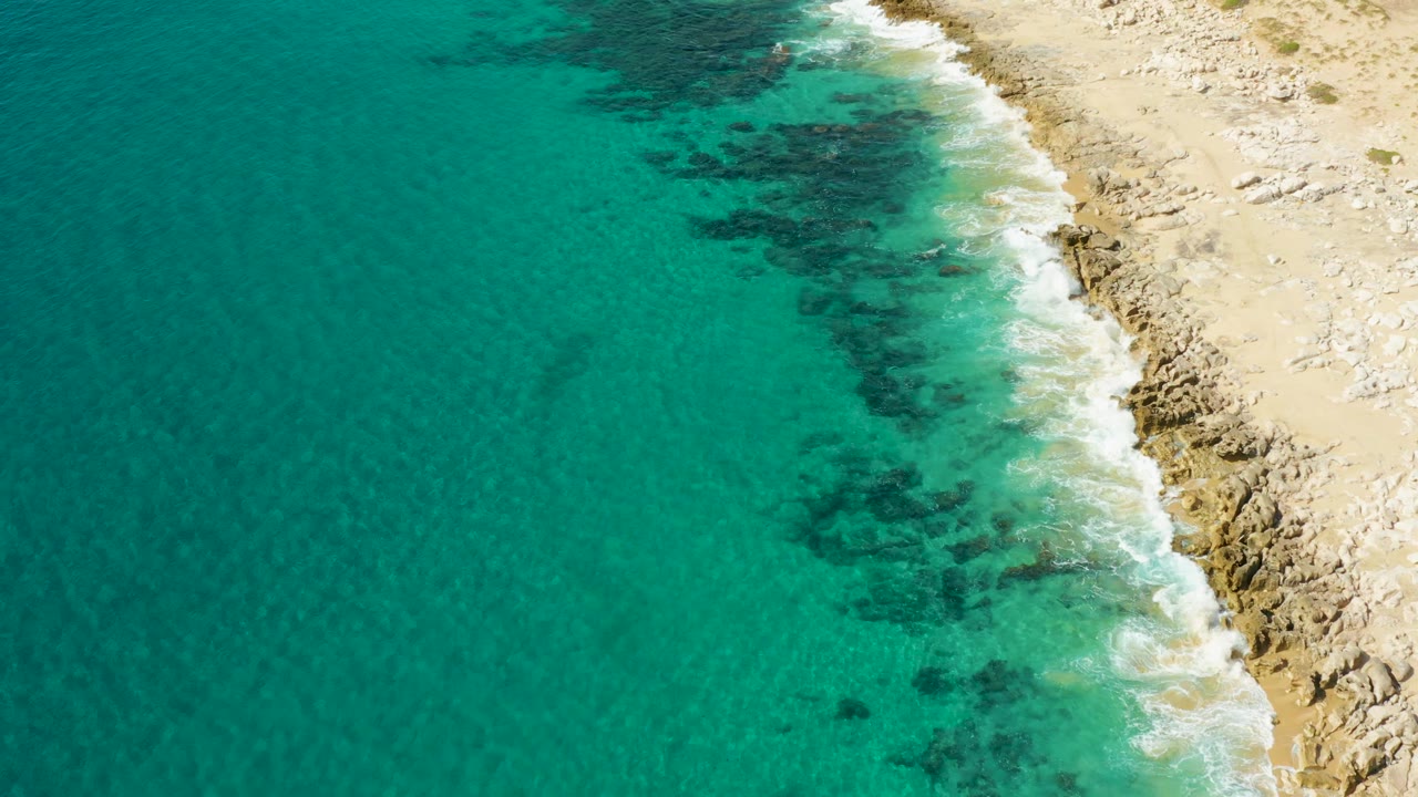 Aerial frame of Laguna Beach Los Cabos shoreline with open turquoise water on the left, foam at the beach edge, rocks on the right, and no visible people.