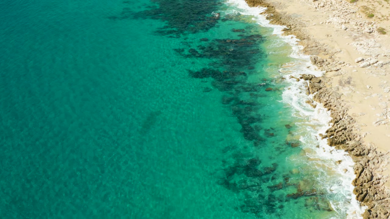 Aerial view over clear turquoise water at Laguna Beach Los Cabos, with the rocky sandy shoreline pressed along the right edge and no visible people.