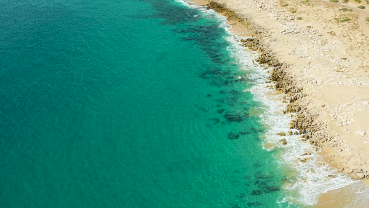 Overhead view of turquoise water meeting the rocky sandy shore at Laguna Beach Los Cabos, with small white waves and no visible people.